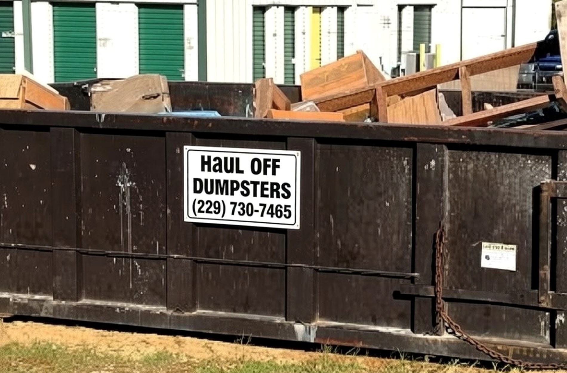 A Haul Off Dumpsters roll-off container loaded with timber and building debris, its white sign plate reading Haul Off Dumpsters and the phone number, parked beside storage units