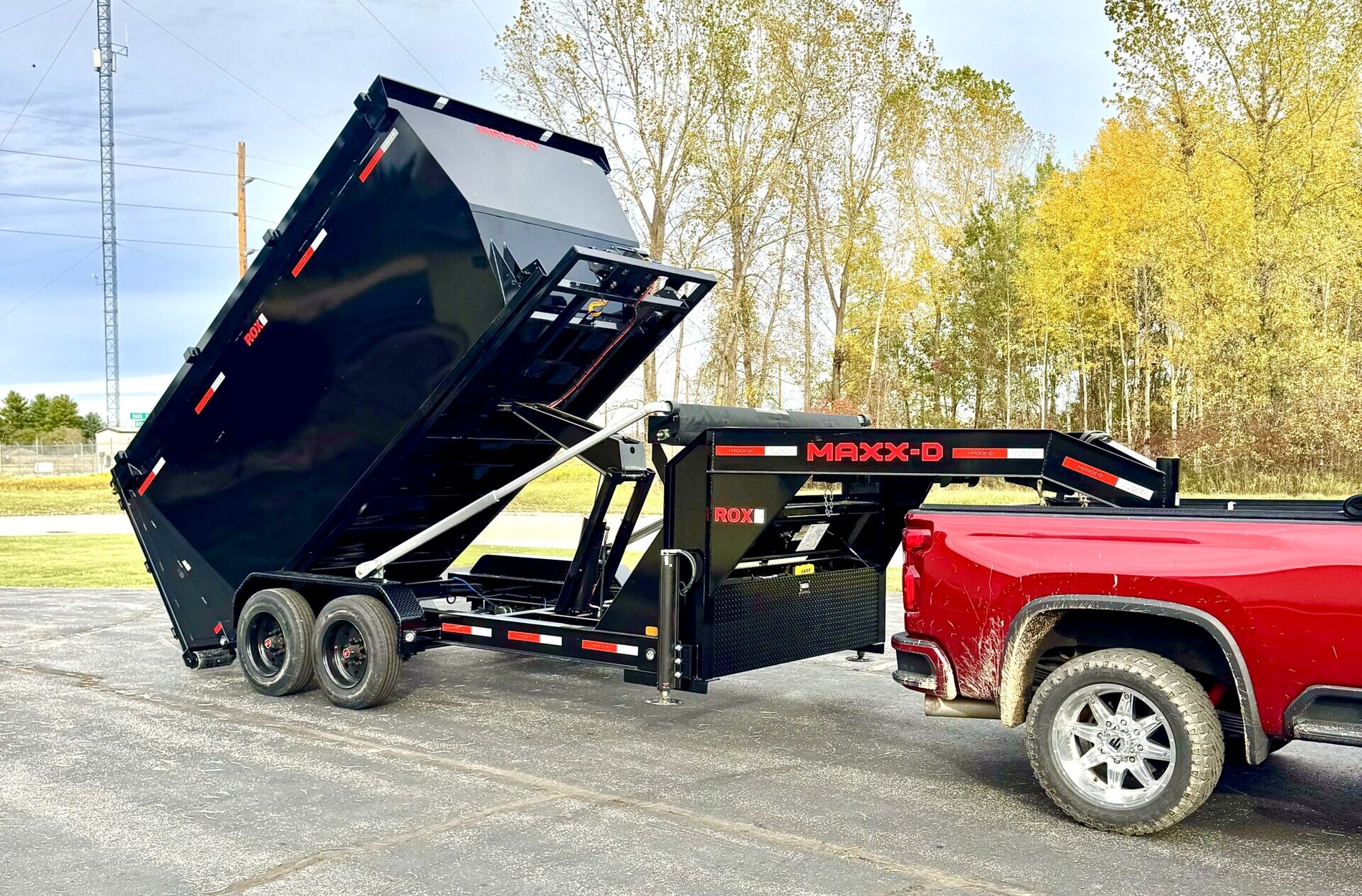 A black gooseneck dump trailer with its body raised to tip a load, hitched to a pickup truck